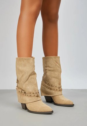 Bottes basses en daim beige décontractées avec des sangles cloutées et des talons carrés bas, portées sur des jambes nues sur un fond uni.