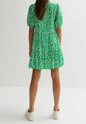 Woman standing wearing a green floral short dress with puff sleeves and white sneakers, facing away against a plain light background.