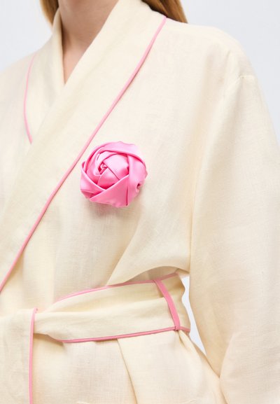 Γυναίκα που φοράει κρεμ ρόμπα με ροζ περιθώριο, δεμένη στη μέση, με ροζ σατέν τριαντάφυλλο καρφιτσωμένο στην αριστερή πλευρά του στήθους.