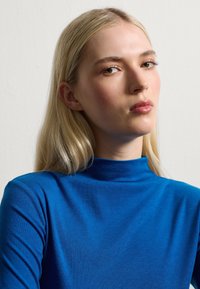Blue, long-sleeve top with a high neck made of smooth, stretchy fabric. The texture appears soft with even coloration and no visible patterns.