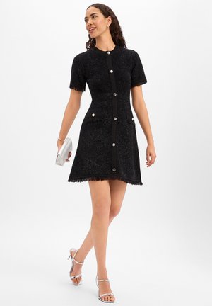 Black textured dress with short sleeves, button front, and fringed hem. Features two front pockets with contrasting black accents.