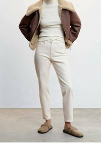 Personne portant un pull côtelé crème, une veste en peau lainée marron, un pantalon blanc cassé et des chaussures beige à enfiler, debout sur un sol en béton.