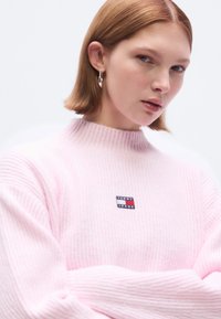 Pulover roz deschis, cu dungi, cu guler înalt, cu o mică aplicație cu logo-ul "TOMMY JEANS" în albastru și roșu pe piept. Textură moale.