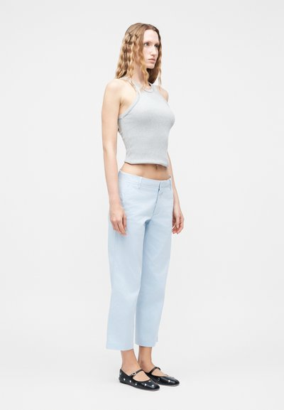 Γυναίκα με μακριά κυματιστά μαλλιά, φορώντας αμάνικο ανοιχτό γκρι τοπ, ανοιχτό μπλε παντελόνι καμπάνες και μαύρα Mary Jane παπούτσια με τρουκς, στέκεται πλάγια.