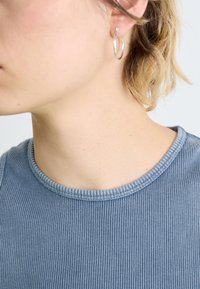 Hõbedane rõngaskõrvarõngas, mida kannab inimene, millel on sile ja läikiv pind. Inimesel on seljas helesinine ribakujuline topp, millel on ümar kaelus.