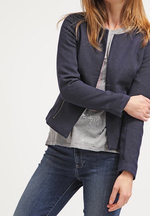 Frau trägt einen marineblauen Blazer mit Reißverschluss über einem grauen T-Shirt mit Grafik und dunkelblauen Jeans, mit einem verschränkten Arm und dem anderen entspannt an der Seite.