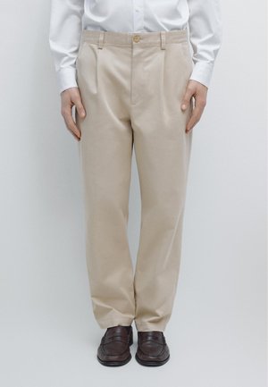 Mann trägt beige Hose, weißes Hemd und dunkelbraune Leder-Loafer, steht mit entspannten Händen seitlich am Körper.