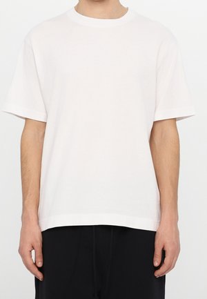 Mann, der ein schlichtes weißes kurzärmeliges T-Shirt mit Rundhalsausschnitt und schwarze Hosen trägt, steht vor einem weißen Hintergrund.