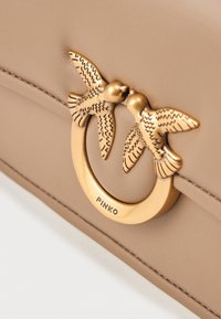 Bolso de mano de cuero beige con un acento de hardware dorado circular, con forma de dos pájaros estilizados, con grabados detallados.