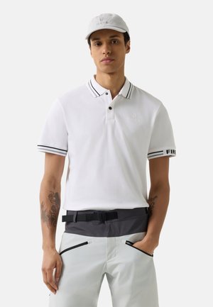 Young man wearing a white polo shirt with striped collar and sleeve edges, white cap, and light-colored pants with black belt and zippered pockets.