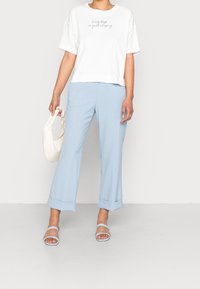T-shirt en coton blanc avec texte, pantalon large bleu clair à revers, et sandales bleu clair. Sac à main beige texturé tenu dans la main gauche.