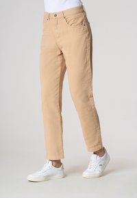 Pantaloni beige chiaro abbinati a sneaker bianche, che mostrano un outfit casual su uno sfondo neutro. Messa a fuoco sulla parte inferiore di un modello in piedi.