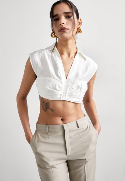 Chemise blanche courte avec col et détails plissés à l'avant, associée à un pantalon taille haute beige clair, au tissu lisse.