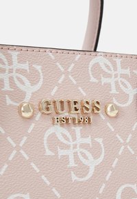 Sac à main synthétique rose clair avec un motif de logo blanc, des accessoires en or et le nom de marque "GUESS EST1981" en relief, agrémenté de clous décoratifs.