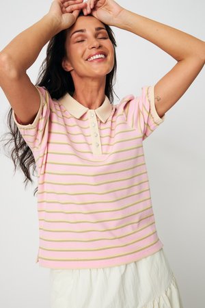 Γυναίκα που χαμογελά με κλειστά μάτια, φορόντας ροζ και κίτρινη ριγωτή πόλο μπλούζα και λευκή φούστα, με τα χέρια σηκωμένα πάνω από το κεφάλι της.