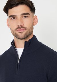 Marineblauwe gebreide trui met een kwartrits, voorzien van een geribbelde kraag, een zilveren rits en een gladde textuur. Gedragen over een witte shirt.