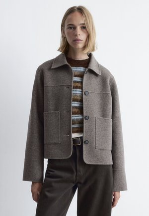 Braune Wollmischungjacke mit Kragen, zwei großen vorderen Taschen und Knopfverschluss. Getragen über einem gestreiften Pullover und dunklen Hosen.