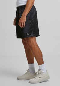 Sorte satin shorts med en glat tekstur, elastisk talje og hvidt logo-detail. Kombinationen er med lyse sneakers og hvide sokker.