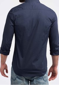 Navyblauwe lange mouw blouse met knopen, opgerolde mouwen, zachte textuur, gebogen zoom en rugpasdetails. Gedragen met lichtblauwe spijkerbroek.