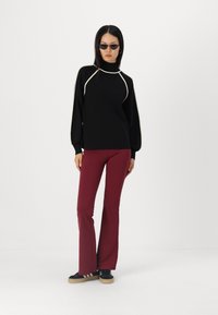 Pull noir avec des bordures blanches, col montant et manches bouffantes, associé à un pantalon évasé bordeaux et des baskets noires avec des rayures blanches.