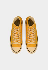 Κίτρινα πάνινα παπούτσια ψηλά με μπεζ κορδόνια, λαστιχένια άκρη δαχτύλου και μαύρη ρίγα κατά μήκος της σόλας. Εμφανές το λογότυπο στις πλευρές.