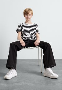 Black and white striped t-shirt, black wide-leg pants, and white high-top sneakers, seated on a minimalist chair against a light background.