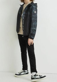 Chaqueta cortavientos de tie-dye en negro y gris con cierre de cremallera, capucha y acento de logo en la manga; combinada con pantalones negros y zapatillas negras con acentos blancos.