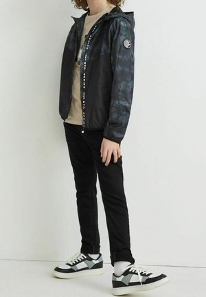 Schwarz-graue Batik-Windbreaker-Jacke mit Reißverschluss, Kapuze und Logo-Akzent am Ärmel; kombiniert mit schwarzen Hosen und schwarzen Sneakers mit weißen Akzenten.