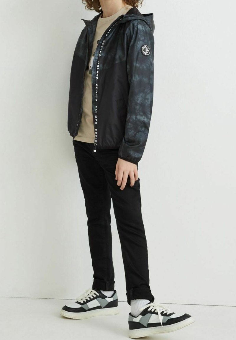 Chaqueta cortavientos de tie-dye en negro y gris con cierre de cremallera, capucha y acento de logo en la manga; combinada con pantalones negros y zapatillas negras con acentos blancos.