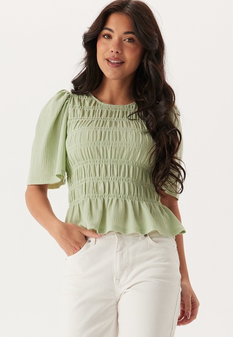 Blusa peplum verde chiaro con maniche a sbuffo, tessuto texturizzato e dettagli smock sul corpetto, abbinata a pantaloni bianchi a vita alta.