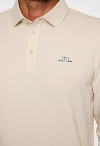 Beige Langarm-Polohemd mit geripptem Kragen, ausgestattet mit einer Knopfleiste mit zwei Knöpfen und einem schwarzen gestickten Logo auf der Brust. Weicher Stoff.