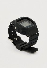 Fekete Casio G-Shock digitális karóra téglalap alakú számlappal, védő lünettával és állítható gumiszíjjal, fehér háttéren.