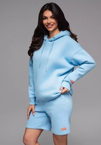 Lichtblauwe sweatshirt met capuchon en voorzak, gecombineerd met bijpassende shorts. Beide gemaakt van zachte stof, met een klein oranje logo.
