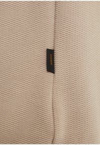 Béžová texturovaná látka s tkaným vzorem a černou etiketou, na které je vytištěno "CHASIN'" oranžovou barvou. Okraj je pečlivě přišitý.