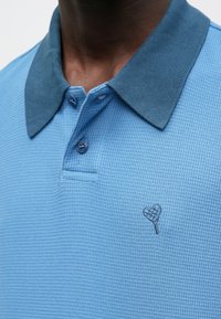 Μπλε πόλο μπλουζάκι με υφή υφάσματος, που διαθέτει γιακά σε πιο σκούρο μπλε χρώμα και μικρό λογότυπο καρδιάς κεντημένο στο στήθος. Με πλάκα δύο κουμπιών.