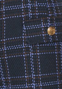 Vlněná tkanina s tmavě námořnickou modrou základnou, s kostkovaným vzorem v světle modré a hnědé, doplněná zlatým knoflíkem na kapse.