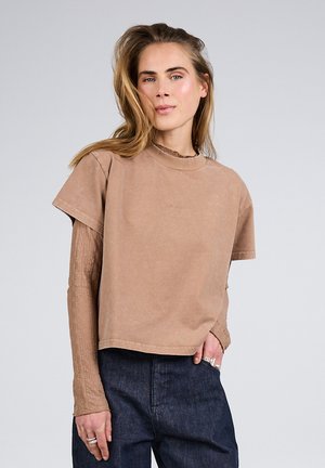 Kvinde med langt hår iført lag-på-lag brun kortærmet og tekstureret langærmet top, mørke jeans og sølvringe, stående foran en grå baggrund.