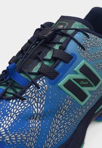 Chaussure de sport bleue et verte avec des motifs texturés réfléchissants, un système de laçage rapide noir et le logo "N Lock" sur le côté.