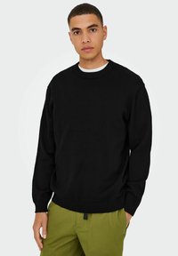 Black sweater with a crew neckline, ribbed cuffs, and hem. Worn over a white shirt, paired with green trousers. Smooth knit texture.