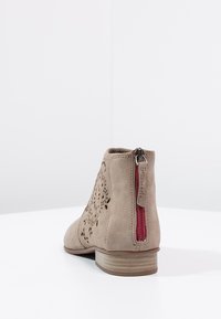 Béžová nubuková kotníková bota s laserem vyřezávaným designem, červeným zipem a nízkým dřevěným podpatkem. Hladká textura, kulatá špička, moderní tvar.