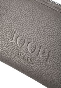 Portafoglio in similpelle grigia con texture e logo "JOOP! JEANS" impresso a rilievo, dotato di chiusura con cerniera argentata. Forma rettangolare compatta.