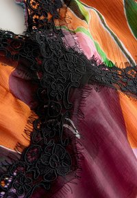 Gros plan d'un tissu multicolore avec des sections orange, rose, verte et violette, orné d'une bordure en dentelle noire florale aux bords effilochés.