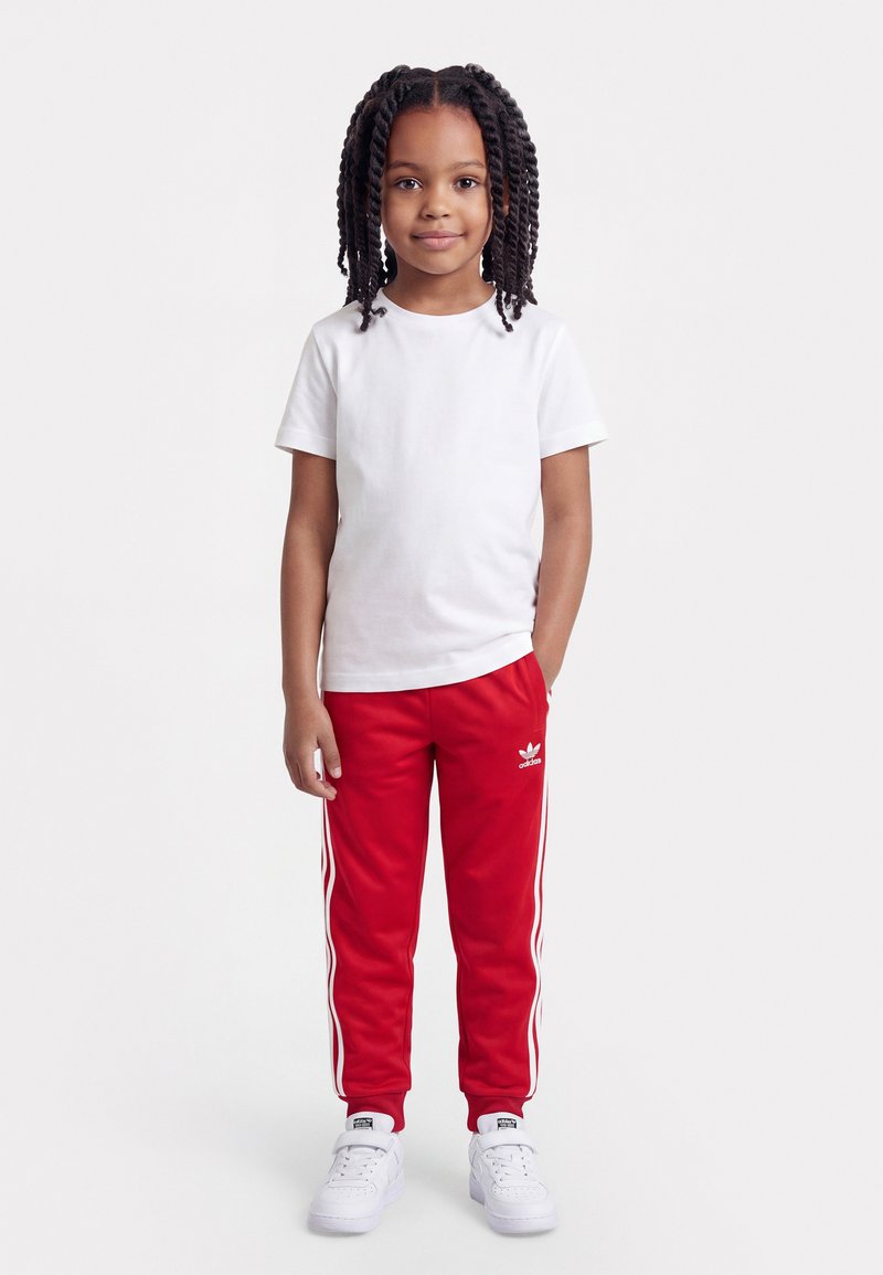 Niña joven con el cabello trenzado, viste una camiseta blanca, pantalones deportivos rojos Adidas con rayas blancas y zapatillas blancas, de pie frente a un fondo blanco.