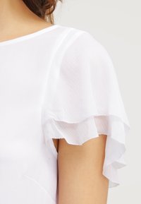 Bílá blůza se průhlednými, volánkovými krátkými rukávy. Hladká tkanina s měkkou texturou, klasický kulatý výstřih a dobře padnoucí střih.