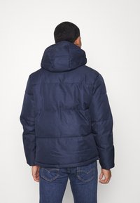 Marinblå vadderad jacka med en strukturerad huva, horisontellt quiltat mönster och en matt yta, bärs över blå jeans.