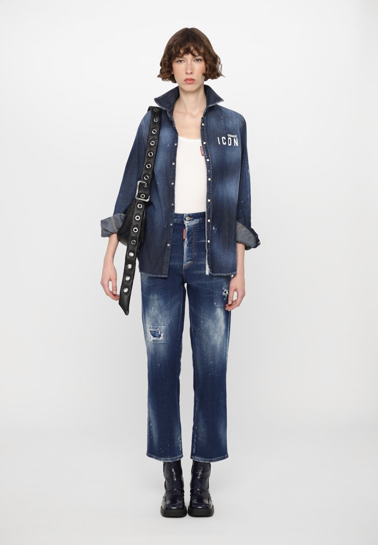 Chemise en denim à boutons avec grand col et texte "ICON", associée à un jean bleu déchiré. Sac utilitaire noir et bottes noires épaisses.