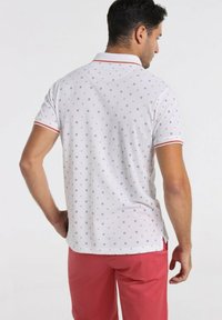 Polo shirt blanc à manches courtes avec de petits motifs colorés, accents rouges sur le col et les manches, associé à un pantalon rouge.