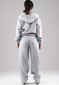 Woman with dark hair in ponytail wearing gray hooded sweatshirt and matching loose sweatpants, standing facing away on plain background.