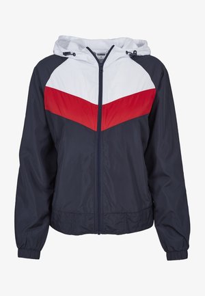 Coupe-vent bleu marine avec une capuche blanche et une bande en chevron rouge sur la poitrine. Comprend une fermeture éclair à l'avant et des poignets élastiques. Tissu léger et texturé.