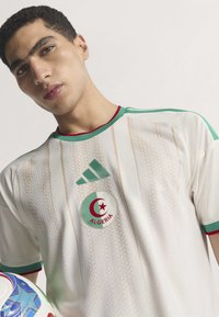 White football jersey with green and red accents. Features the Adidas logo and Algeria crest. Textured fabric with vertical stripes.